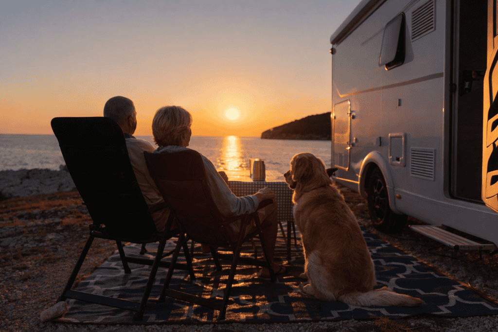 Twee ouderen genieten van een rustige camperplek aan zee bij zonsondergang, met hun hond en camper, perfect voor 60-plussers die willen ontspannen tijdens hun camperreis in Europa.